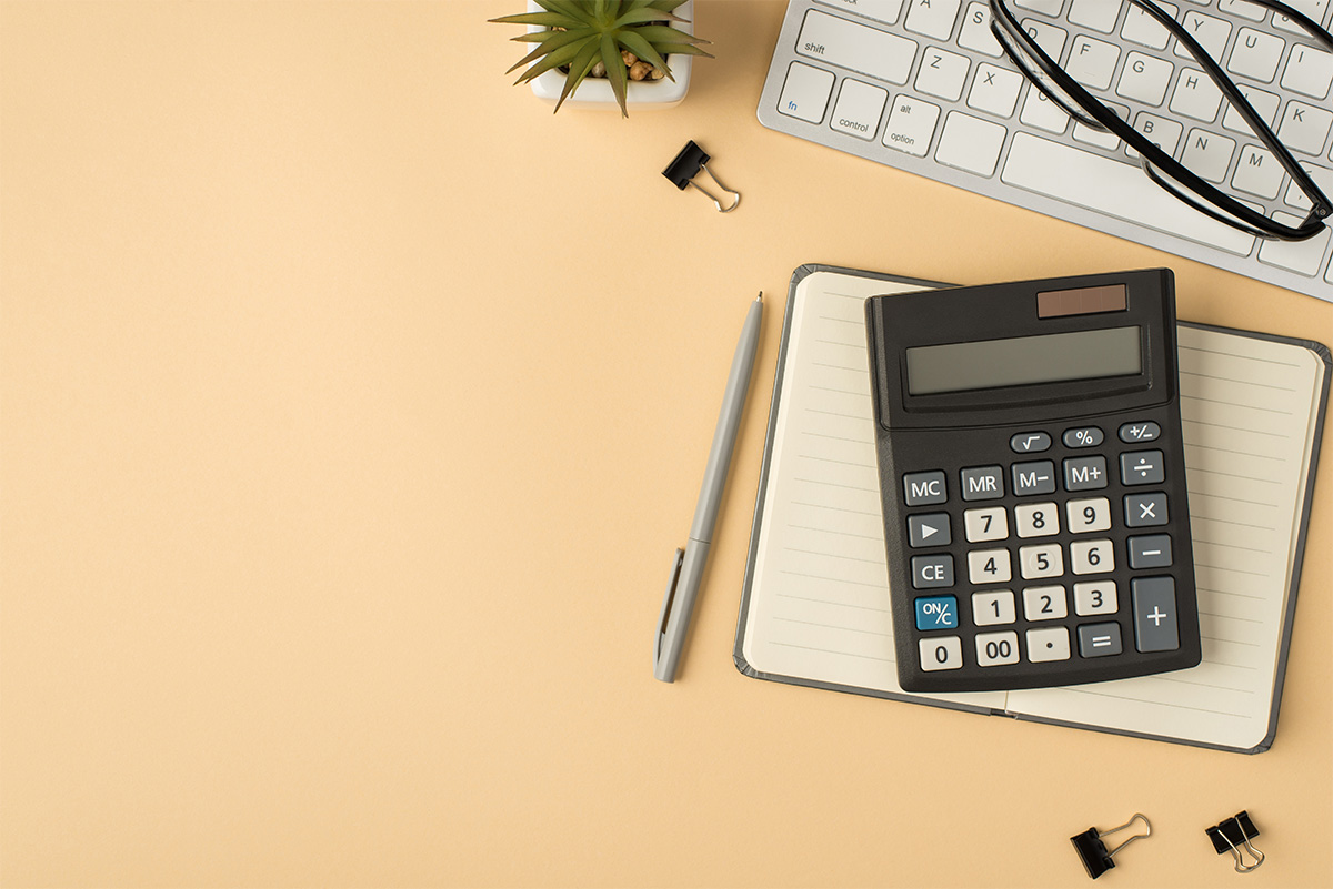 ベージュの背景の上に、ノート、電卓、ペン、眼鏡、キーボード、植物、クリップが置かれたデスクトップの様子。