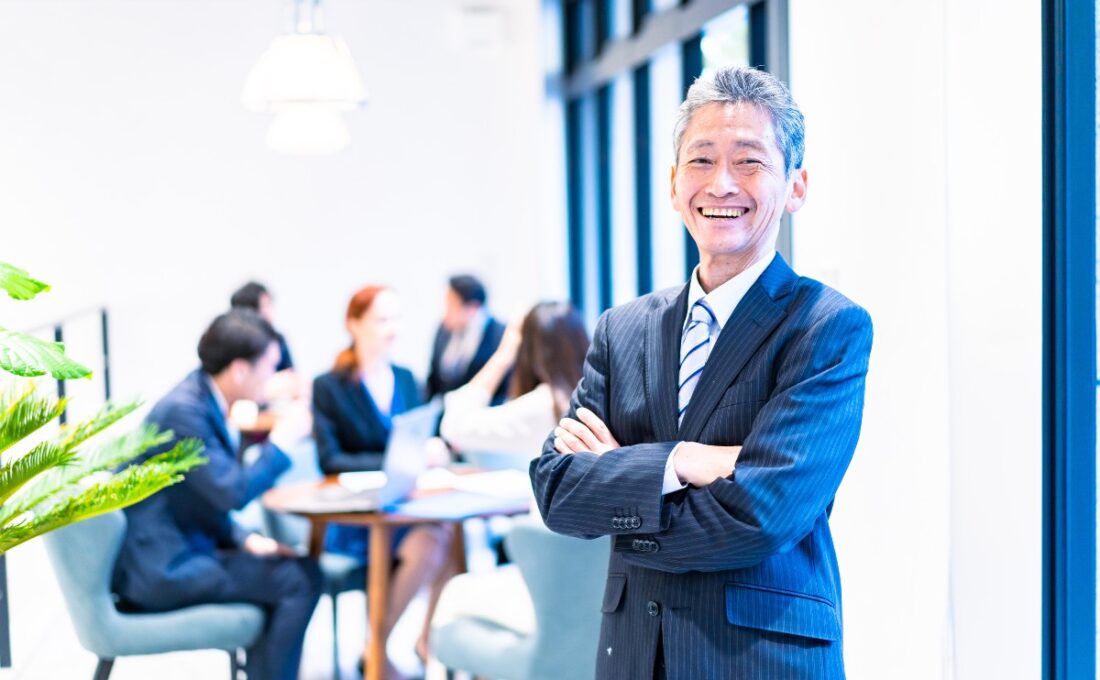 「明るいオフィスで、スーツを着たシニアビジネスマンが腕を組んで笑顔で立っている。背景にはビジネスミーティングをしている同僚たちが映っており、リーダーシップや自信を象徴する場面。」