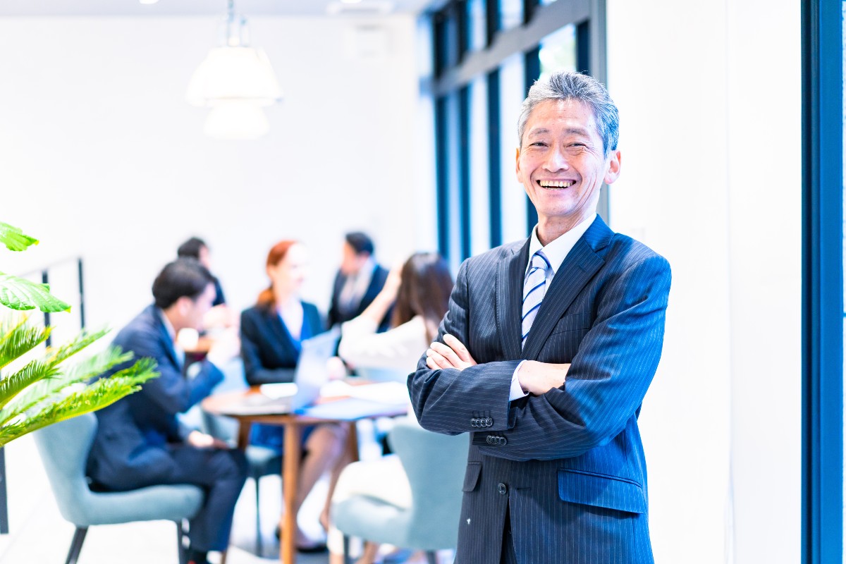 「明るいオフィスで、スーツを着たシニアビジネスマンが腕を組んで笑顔で立っている。背景にはビジネスミーティングをしている同僚たちが映っており、リーダーシップや自信を象徴する場面。」