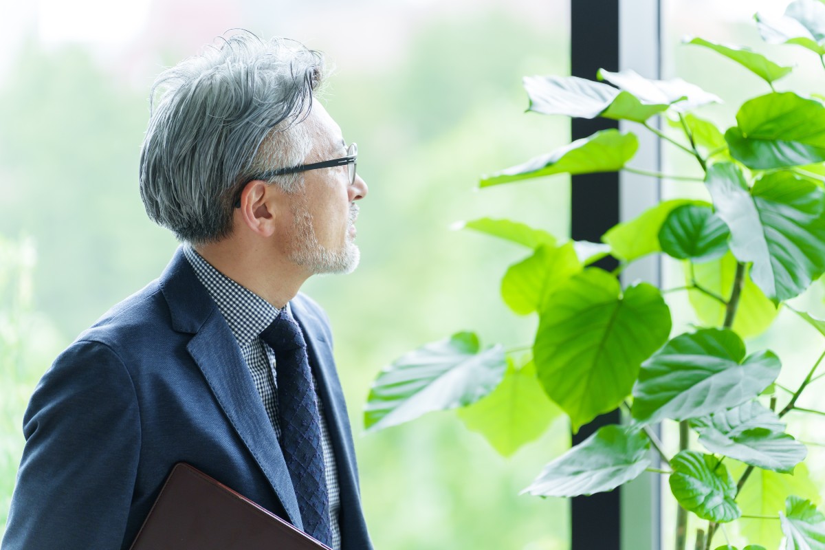 「窓辺に立ち、外を見つめるシニアビジネスマン。緑の植物がそばにあり、リラックスした雰囲気で未来を考えている様子を表している。」