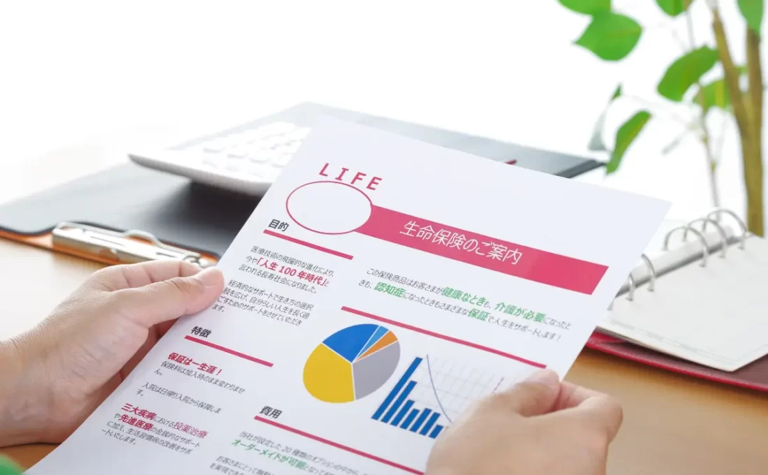 「生命保険の案内書を手に取って読んでいる人物の様子。カラフルなグラフや説明文が書かれており、生命保険の内容について検討している場面。」