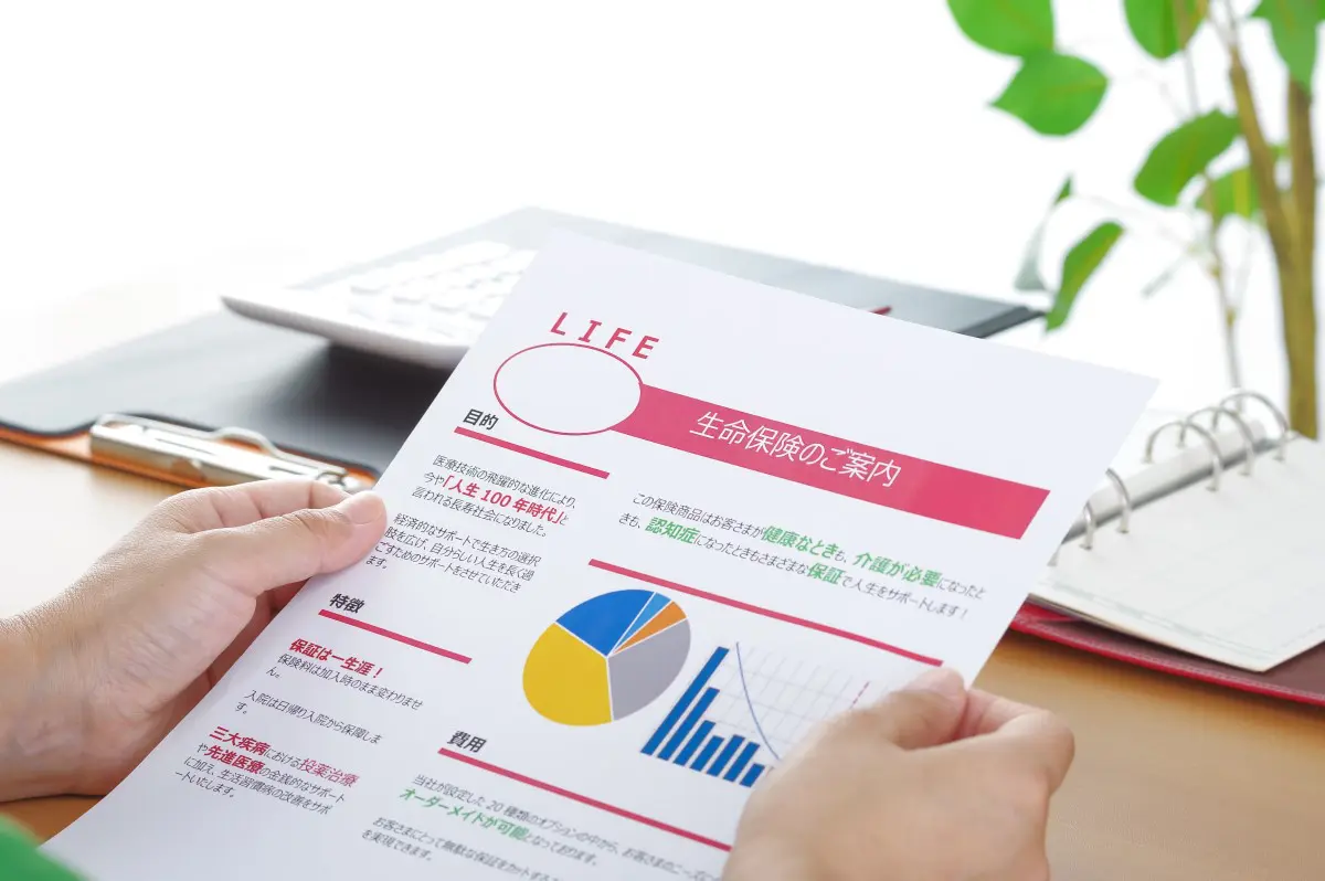 「生命保険の案内書を手に取って読んでいる人物の様子。カラフルなグラフや説明文が書かれており、生命保険の内容について検討している場面。」