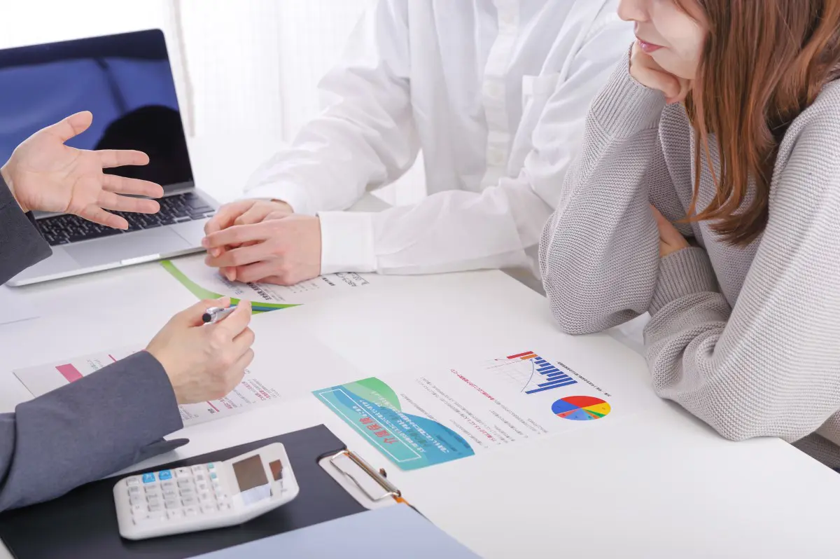 会議テーブルで、複数のビジネスパーソンが資料を見ながら話し合っている様子。グラフや計算機がテーブルに置かれており、ビジネスプランや投資に関する話し合いが行われていることを示している。