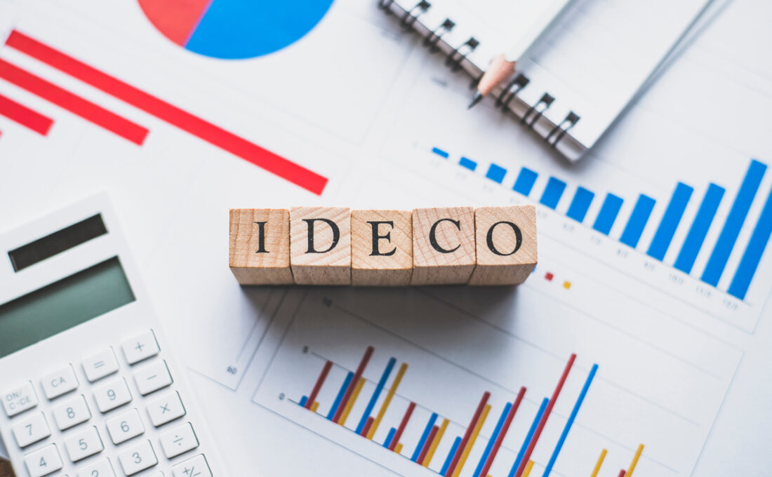 iDeCo（個人型確定拠出年金）に関連するグラフやデータが載った書類の上に、「iDeCo」という文字が書かれた木製ブロックが置かれている。