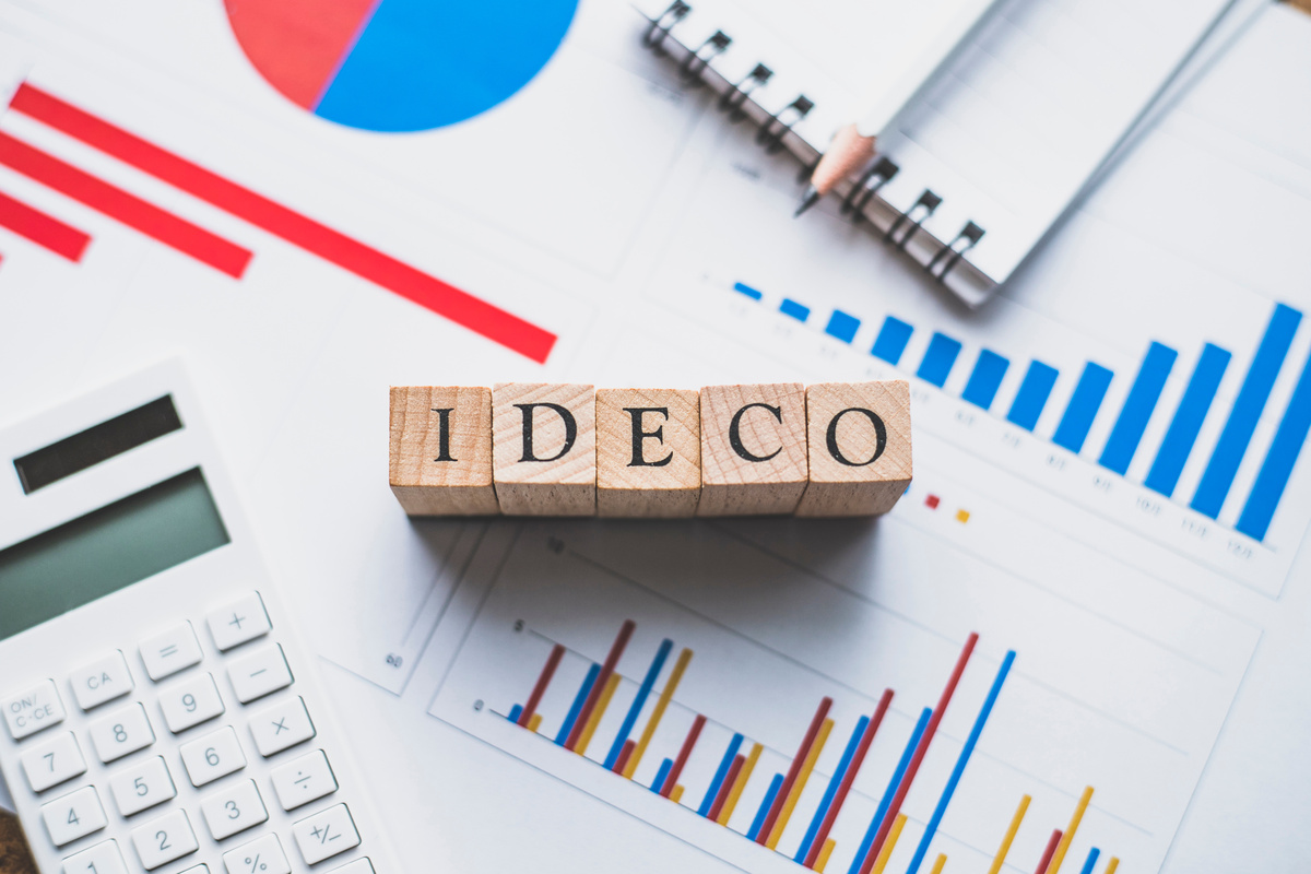iDeCo（個人型確定拠出年金）に関連するグラフやデータが載った書類の上に、「iDeCo」という文字が書かれた木製ブロックが置かれている。