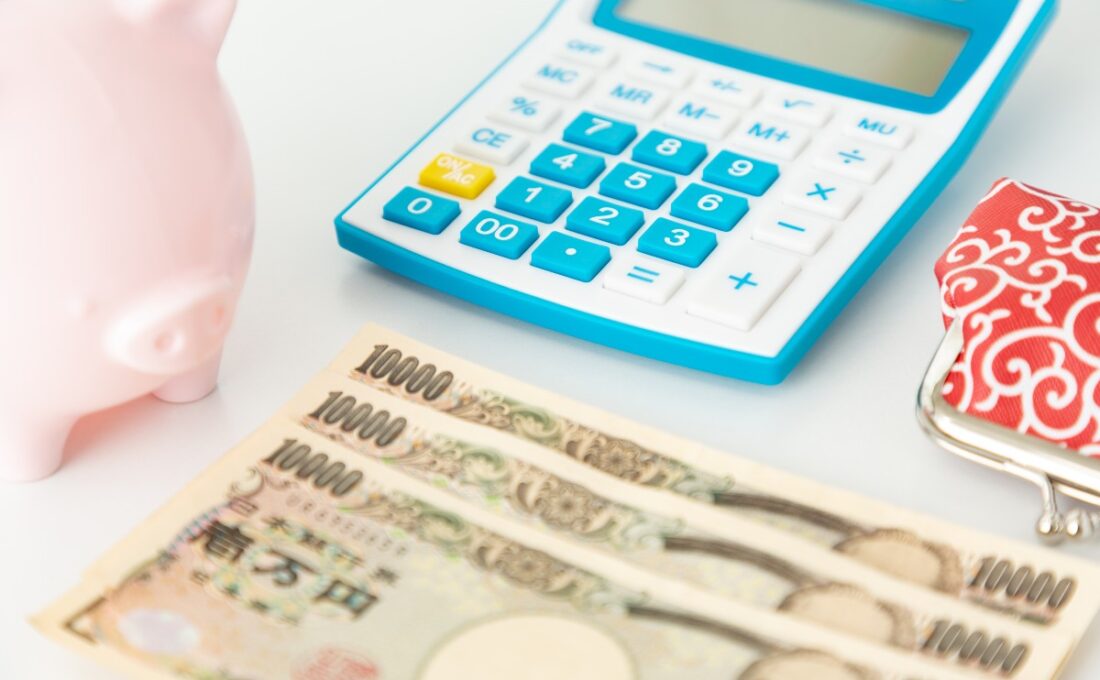 青い電卓と1万円札、ピンクの貯金箱が置かれた机の上のイメージ