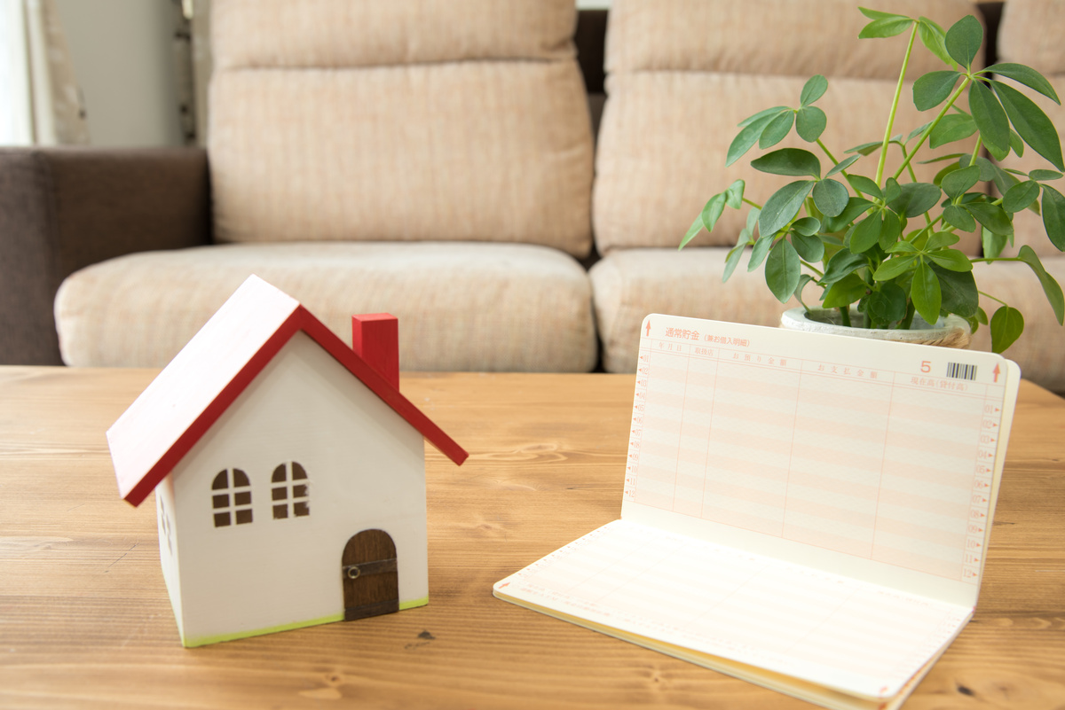 家の模型、銀行通帳、観葉植物が木製のテーブルの上に置かれたリビングの風景