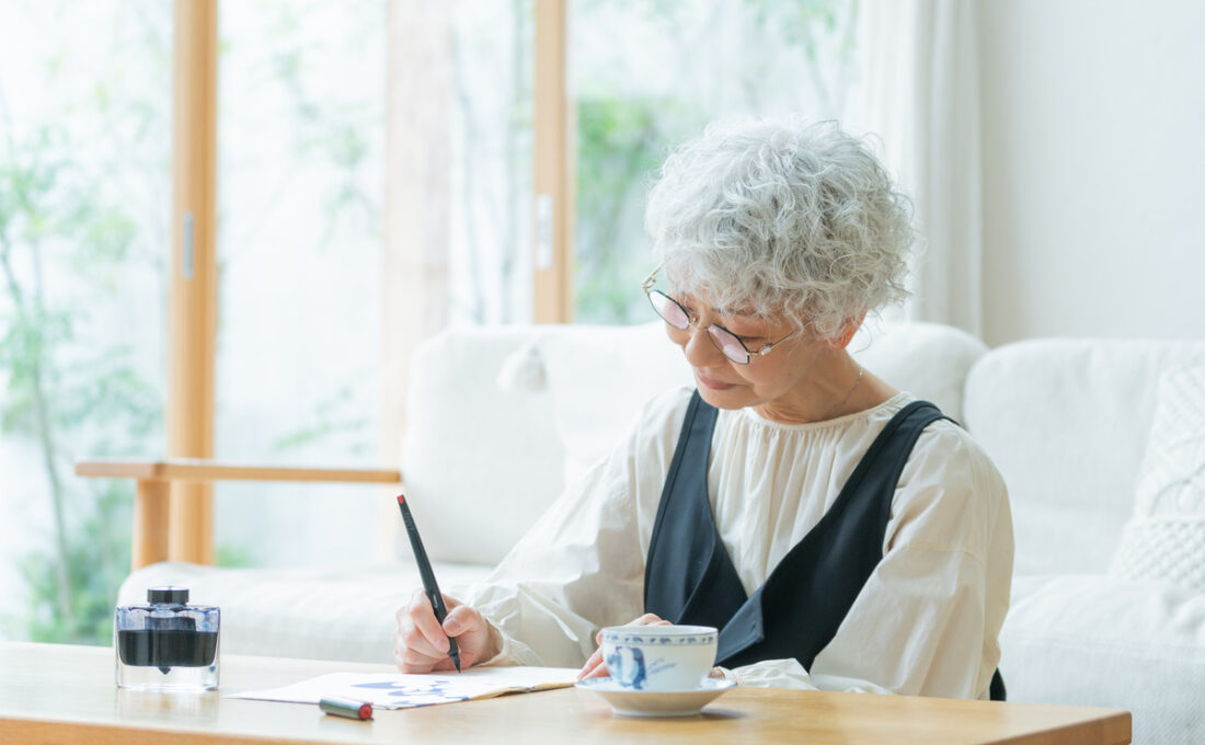 眼鏡をかけた高齢女性が、ペンを使ってノートに書き物をしている。