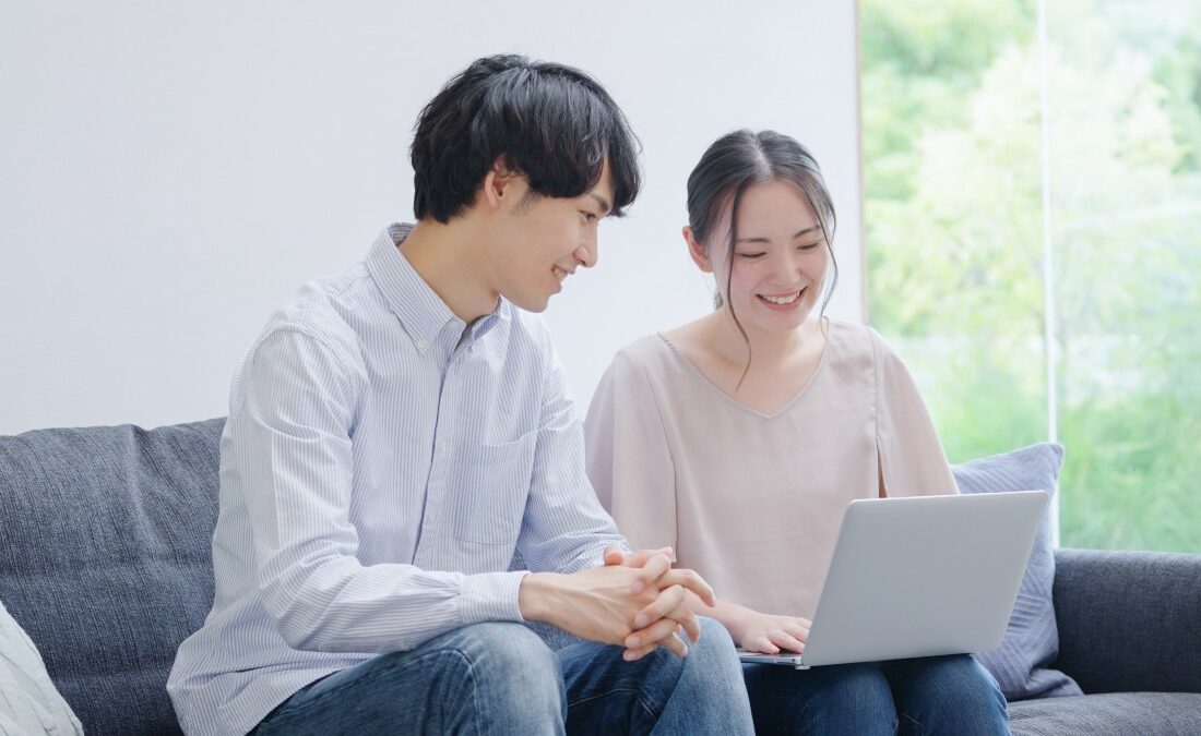 リビングで一緒にノートパソコンを見ながら笑顔で話す若い夫婦