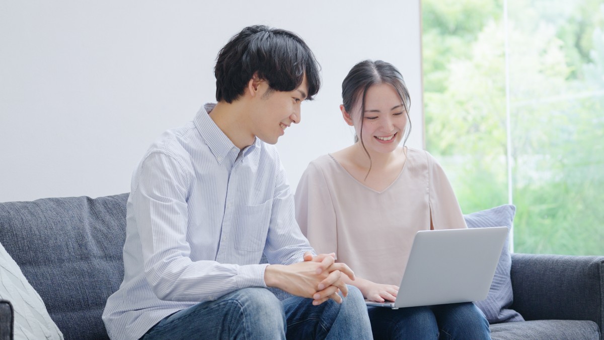 リビングで一緒にノートパソコンを見ながら笑顔で話す若い夫婦