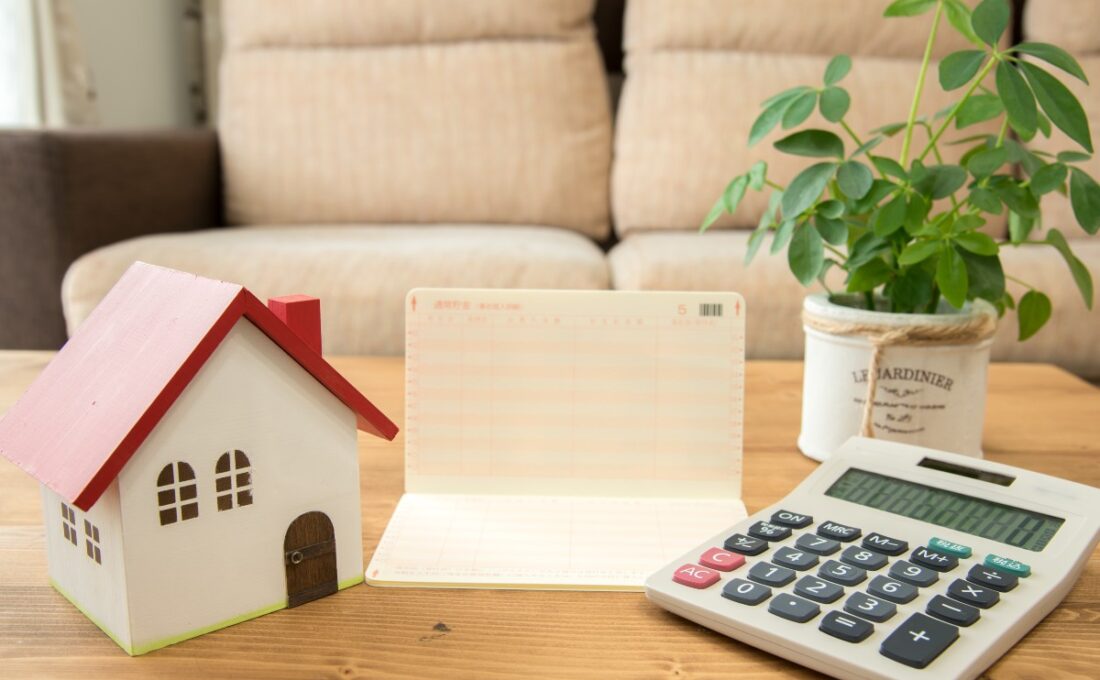 家の模型、電卓、通帳、観葉植物が木製のテーブルに置かれたリビングの風景