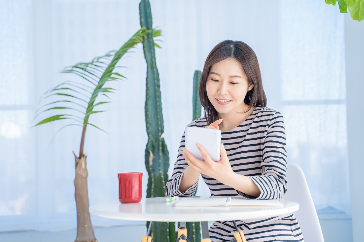 ストライプの服を着た女性がタブレットを使ってテーブルに座り、後ろには観葉植物と赤いマグカップがある