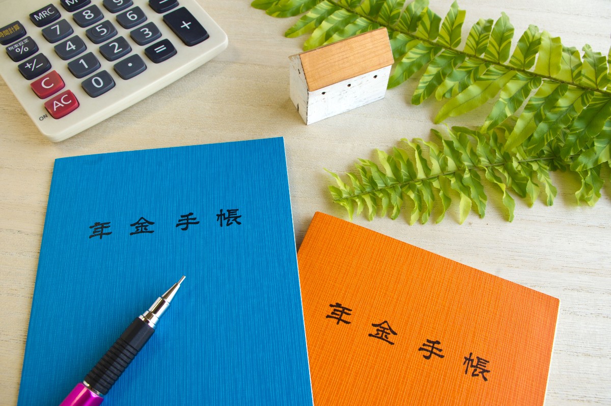 年金手帳、電卓、ペン、小さな家の模型と植物がデスク上に置かれている。