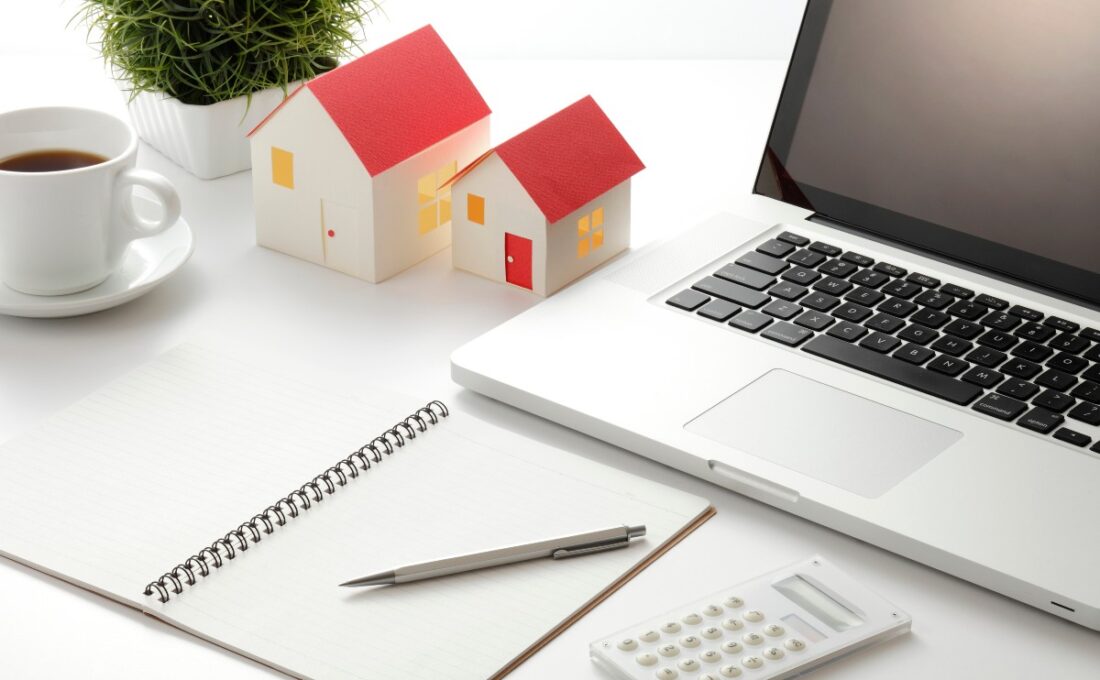 ノートパソコン、家の模型、ノート、電卓、ペン、コーヒーカップが置かれたデスクの風景