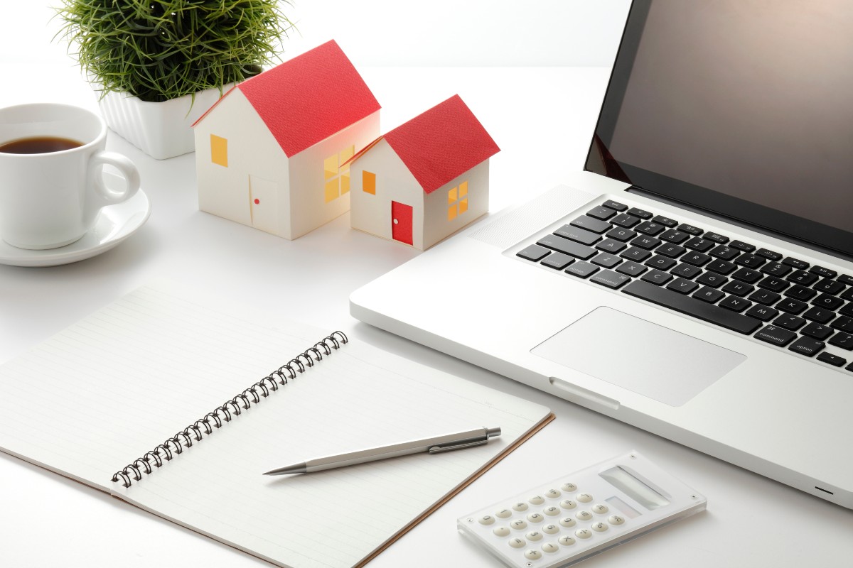 ノートパソコン、家の模型、ノート、電卓、ペン、コーヒーカップが置かれたデスクの風景