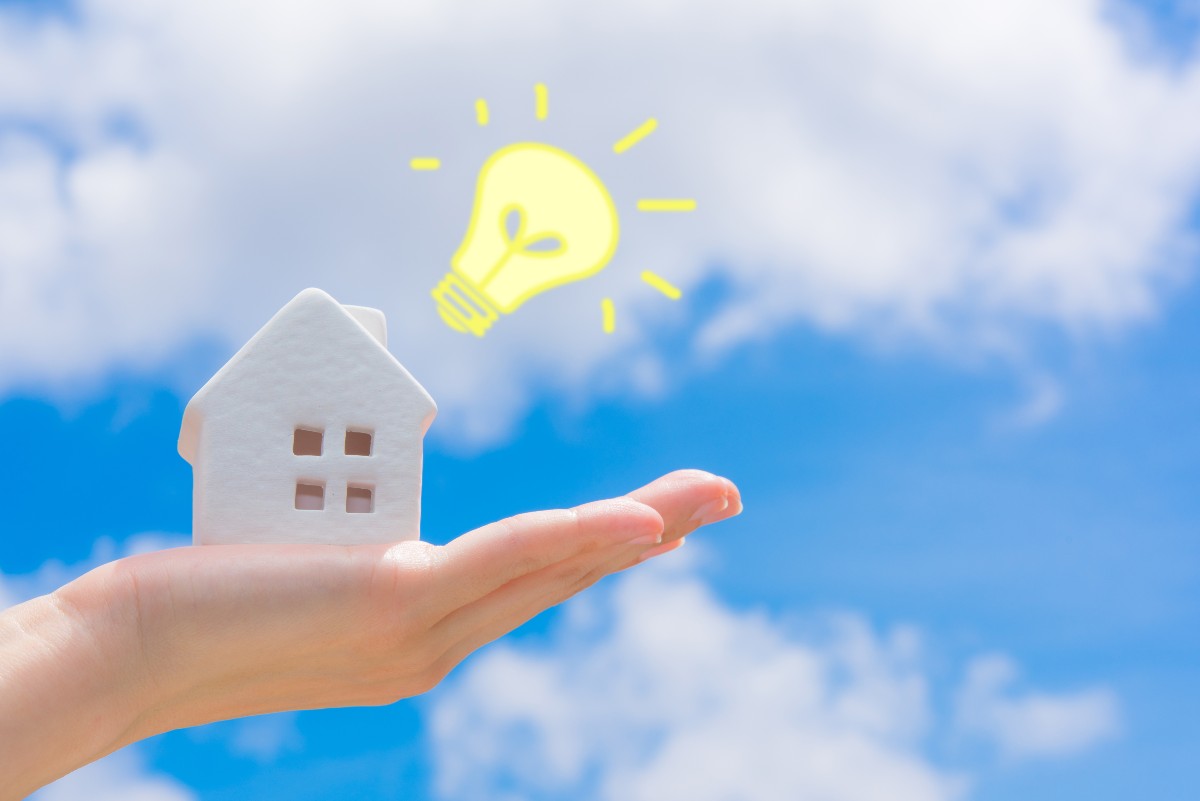 手のひらに乗った家の模型と空に浮かぶ電球のイメージ