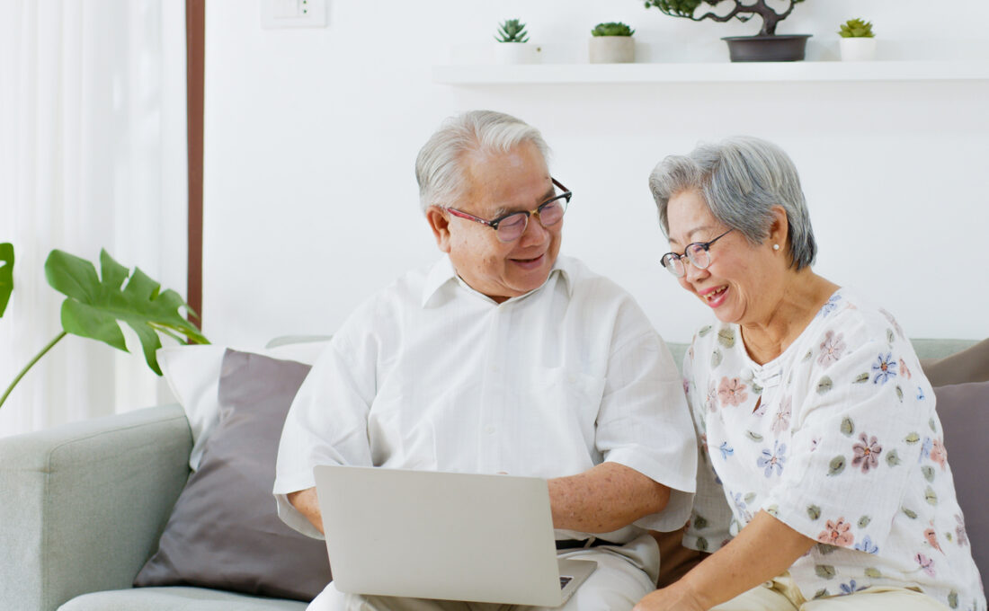 ソファに座って一緒にノートパソコンを見ながら微笑む年配の夫婦