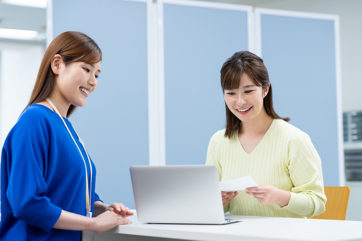 二人の女性がパソコンと書類を見ながら話し合っている様子